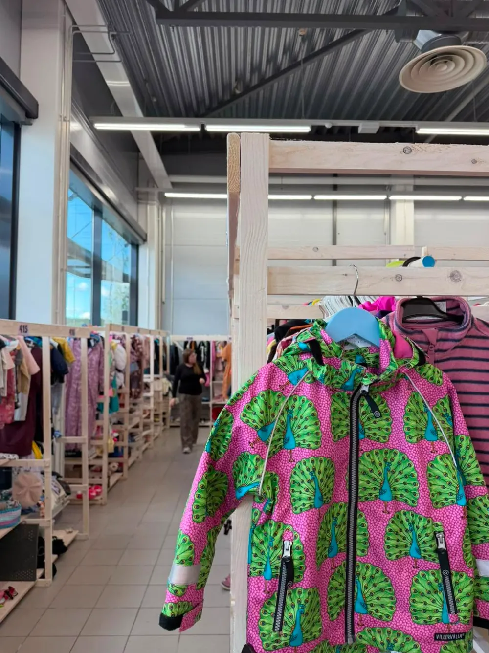Pinkki riikinkukkokuvioinen lasten ulkoiluasu henkarissa, taustalla lastenvaatteiden rivistöjä valoisassa hallissa.